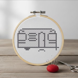 May include: A cross-stitch embroidery hoop featuring a camper design in shades of gray and black. A red pin cushion with needles and small scissors are placed on a wooden surface, suggesting a crafting theme.