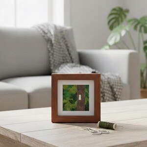 May include: A framed cross-stitch artwork featuring a green and brown landscape design. The wooden frame surrounds the artwork, which is displayed on a light-colored wooden table. A spool of green thread and small scissors are also on the table.