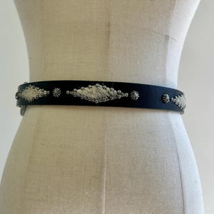 Puede incluir: Un cinturón de cuero negro con adornos de metal plateado. El cinturón presenta elementos decorativos en forma de diamante y estrella con un relleno blanco texturizado. El cinturón se muestra en un maniquí.