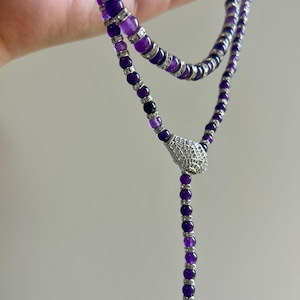May include: A layered necklace featuring strands of deep purple beads and silver accents. The necklace has a decorative silver pendant with clear stones. The beads are a mix of sizes, creating a textured look. The necklace is designed to be worn around the neck.