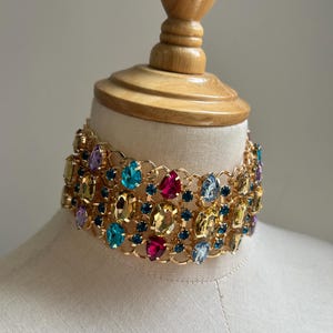 May include: A gold choker necklace with a vibrant gemstone arrangement. The necklace showcases a variety of oval-cut gemstones in shades of blue, yellow, pink, and purple. The gems are set in a gold-coloured metal frame.
