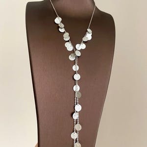 Puede incluir: Un collar plateado con un diseño en forma de Y. El collar presenta múltiples colgantes pequeños, redondos y con forma de moneda que caen en cascada por la parte delantera. Los colgantes están espaciados uniformemente y conectados por delicadas cadenas. El collar se exhibe en un expositor de joyas marrón.