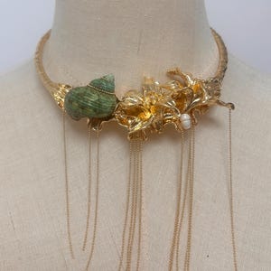 May include: A gold-toned metal choker necklace featuring a textured band, a green spiral seashell, and metallic floral accents. The piece is adorned with a single small white pearl and multiple delicate gold chains that hang down from the center.