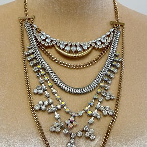 May include: A multi-layered statement necklace featuring gold-tone chains, a silver-tone snake chain, and clear crystal accents. The design includes a crescent-shaped gold element, floral-inspired crystal clusters, and iridescent stones.