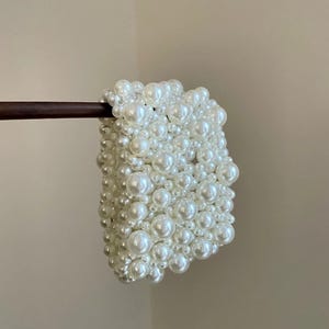 May include: A white pearl beaded clutch bag. The bag is rectangular and densely covered with various sizes of white pearls. It is held by a thin, dark brown wooden handle. The background is a neutral beige color.
