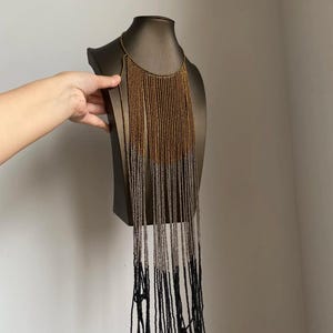 May include: A beaded necklace with a cascading design. The necklace features a gradient of colors, starting with gold at the top, transitioning to silver, and ending with black beads. The necklace is displayed on a dark brown mannequin.