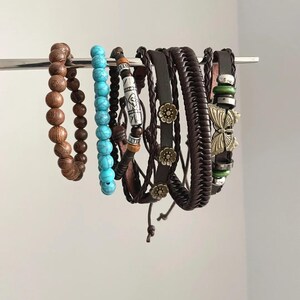 Puede incluir: Una colección de pulseras de varios estilos y materiales. Las pulseras incluyen cuentas de madera, cuentas turquesas, cuero y diseños trenzados. Algunas presentan elementos decorativos como flores metálicas y una mariposa.