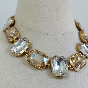May include: A statement necklace featuring large, rectangular, faceted crystals in alternating clear and champagne-colored tones. Each crystal is set in a gold-toned metal frame, linked together to form a continuous collar-style piece of jewelry.