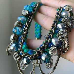 Könnte beinhalten: Ein geflochtenes Armband aus ineinander verschlungenen blauen und grünen Stoffsträngen, verziert mit facettierten klaren und blauen Kristallperlen in Metallfassungen sowie mehreren dünnen Ketten in antikem Messington.