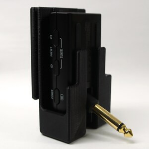 Op de afbeelding: Een zwart elektronisch apparaat met een goudkleurige audio jack, gehouden in een zwarte beugel. Het apparaat heeft knoppen met de labels "Preset", "Tuner" en "Power". De jack is schuin geplaatst.