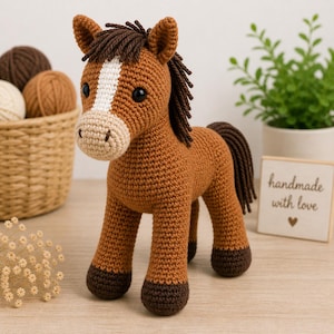 Puede incluir: Caballo de ganchillo hecho a mano en tonos marrón, beige y blanco. El caballo tiene una raya blanca en la cara, crin y cola marrón oscuro y cascos marrones oscuros. Se encuentra sobre una superficie clara.