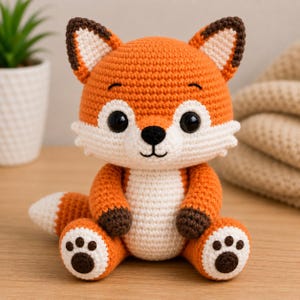 Könnte beinhalten: Ein handgehäkelter Fuchskuscheltier in Orange, Weiß und Braun. Der Fuchs hat große schwarze Augen, eine weiße Brust und einen flauschigen Schwanz. Er sitzt auf einer Holzoberfläche.