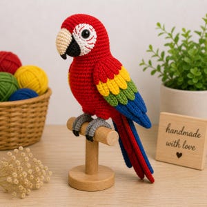 May include: A handmade, crochet parrot in vibrant red, yellow, green, and blue. The bird is perched on a wooden stand, with a beige beak and detailed eye. A wooden sign with "handmade with love" is visible.