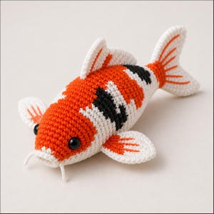 Op de afbeelding: Een gehaakt speelgoed van een koikarper in oranje, wit en zwart. De vis heeft zwarte kraaloogjes, witte vinnen met oranje accenten en een witte mond met twee kleine snorharen. Op een witte ondergrond.