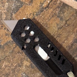 May include: A black utility knife with a silver blade and a white sliding mechanism. The knife has a textured handle with several hexagonal and rectangular cutouts. A green and tan cord is attached to the end of the handle.
