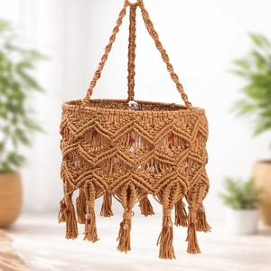Puede incluir: Lámpara colgante de macramé marrón claro, con intrincados patrones de nudos y borlas decorativas. Pequeñas cuentas rojas y transparentes adornan los flecos. La lámpara se suspende de una cadena metálica, ideal para decoración bohemia.