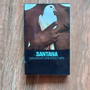 May include: A vintage cassette tape of Santana's Greatest Hits. The album cover features a white dove held by a person with brown skin. The band's name is in turquoise, with the album title below in white text.