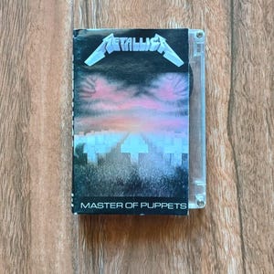 Puede incluir: Una cinta de casete del álbum "Master of Puppets" de Metallica. La portada presenta el logotipo de la banda en plata y una escena de cementerio con un cielo rosa y púrpura. El casete está en una caja de plástico transparente.