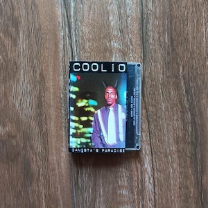May include: A vintage cassette tape featuring the album "Gangsta's Paradise" by Coolio. The album cover shows Coolio in a purple shirt and white tie. The cassette is on a wooden surface.