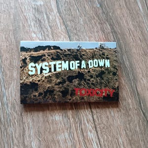 May include: A rectangular magnet featuring the words "SYSTEM OF A DOWN" in white block letters, above the word "TOXICITY" in red. The background depicts a brown hillside with a blue sky.