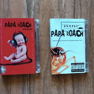 Może przedstawiać: Dwa etui na płyty CD z zespołem Papa Roach. Jedno etui jest czerwone z dzieckiem w słuchawkach, drugie białe z grafiką karalucha. Oba etui zawierają nazwę zespołu i tytuły albumów.