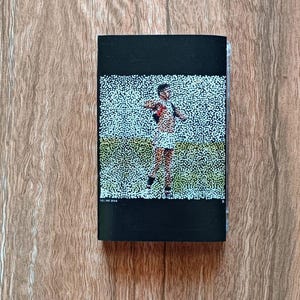 Puede incluir: Un objeto rectangular negro con una imagen punteada de una persona con ropa deportiva en un campo verde. La persona lleva un uniforme rojo y blanco. El fondo es una veta de madera marrón claro.