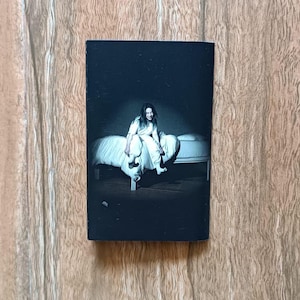 Op de afbeelding: Een zwart-witfoto van een persoon die op een bed zit. De persoon draagt witte kleding en glimlacht. Het bed heeft wit beddengoed. De achtergrond is donker, met een schijnwerper op de persoon.