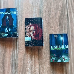 Puede incluir: Tres estuches de CD sobre una superficie de madera. Los estuches presentan portadas de álbumes de "Encore", un retrato y "Curtain Call" de Eminem. El estuche "Encore" muestra una figura en un escenario. El estuche "Curtain Call" tiene el título del álbum.