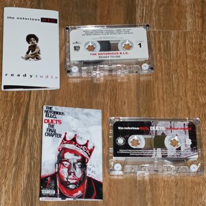 May include: Four items are displayed on a wooden surface: two cassette tapes and two album covers. The album covers feature the text "the notorious B.I.G." and "ready to die" and "Duets The Final Chapter". The cassette tapes are clear plastic.