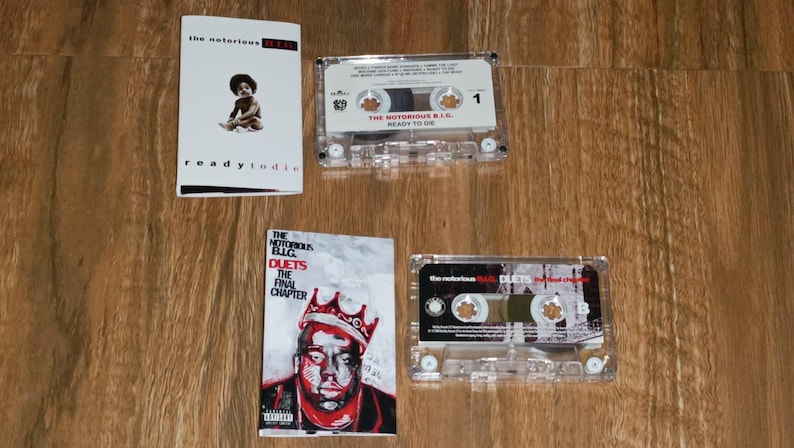 May include: Four items are displayed on a wooden surface: two cassette tapes and two album covers. The album covers feature the text "the notorious B.I.G." and "ready to die" and "Duets The Final Chapter". The cassette tapes are clear plastic.