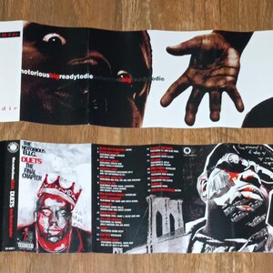 May include: Folded CD case featuring album art for "Ready to Die" and "Duets: The Final Chapter" by The Notorious B.I.G. The artwork includes portraits, a baby, and the Brooklyn Bridge. The color palette is primarily black, white, and red.