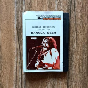 May include: A vintage album cover for George Harrison's Concert for Bangla Desh. The cover features a photo of Harrison singing into a microphone. The album title and artist's name are printed in a bold, sans-serif font. The Aquarius logo is at the top.