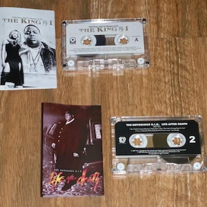 May include: Two cassette tapes and two album covers are displayed on a wooden surface. The cassette tapes are clear with black labels. The album covers feature images of the artist. The text "THE KING & I" and "LIFE AFTER DEATH" are visible.