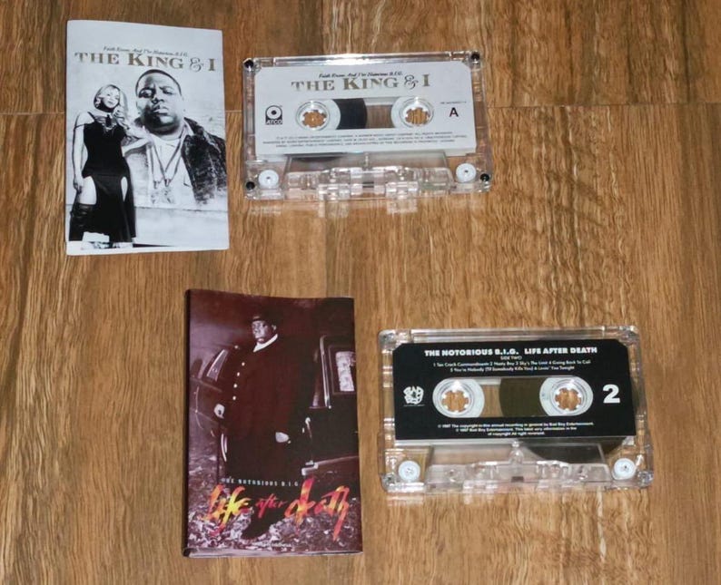 May include: Two cassette tapes and two album covers are displayed on a wooden surface. The cassette tapes are clear with black labels. The album covers feature images of the artist. The text "THE KING & I" and "LIFE AFTER DEATH" are visible.