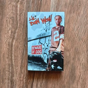 以下が含まれることがあります： 長方形のLil' Bow Wow CDケース。背番号2のフットボールジャージを着た若い人が、大きな黒い犬と一緒に立っています。「BEWARE OF DOG」の文字とアーティスト名が見えます。