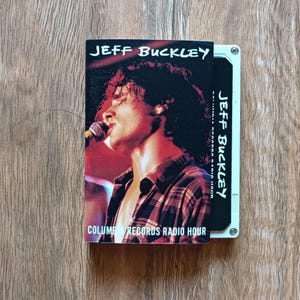 May include: A cassette tape featuring Jeff Buckley. The cassette cover displays a photo of Buckley singing into a microphone. The text "JEFF BUCKLEY" is at the top, and "COLUMBIA RECORDS RADIO HOUR" is at the bottom. The cassette is on a wooden surface.
