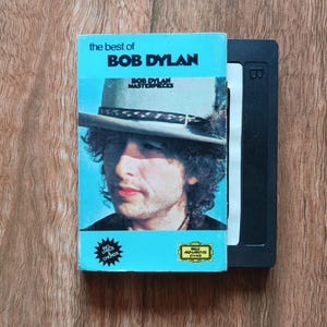 May include: A vintage VHS tape of "The Best of Bob Dylan" with a light blue cover featuring a portrait of Bob Dylan. The tape is next to a black VHS case. The cover text includes "Bob Dylan Masterpieces".