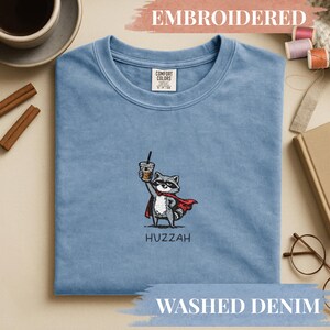 Könnte beinhalten: Hellblaues Washed-Denim-T-Shirt mit einem gestickten Waschbären, der einen Umhang trägt und eine Kaffeetasse hält. Das Wort "Huzzah" ist darunter gestickt.