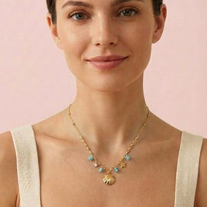 Puede incluir: Un collar dorado con un colgante de concha y pequeñas cuentas turquesas. El collar tiene una cadena delicada e incluye un dije de estrella. La mujer lleva una camiseta sin mangas blanca.