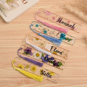 May include: Clear resin bookmarks with dried flowers and names: Hannah, Chloe, Elara, Olivia, and Stella. Each bookmark has a tassel. A book and flowers are in the background.