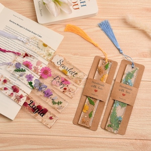 Puede incluir: Una colección de marcapáginas artesanales de resina con flores secas y borlas. Algunos marcapáginas tienen nombres como Scarlett, Ruby, Amelia y Layla. Dos marcapáginas están unidos a tarjetas marrones con el texto "Handmade with love".