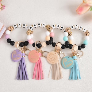 Peut inclure: Quatre bracelets porte-clés perlés avec le mot "MAMA" épelé en perles blanches et imprimé léopard. Chaque bracelet a un pompon et un charme de couleur différente avec un cœur ou le mot "MAMA".