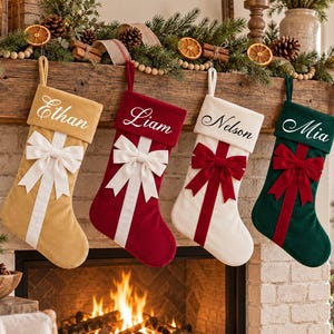以下が含まれることがあります： ベルベット製のクリスマスストッキングが4つ並んで吊るされています。左から順に、白いリボンの付いたゴールド、白いリボンの付いた赤、赤いリボンの付いた白、赤いリボンの付いた緑のストッキングです。それぞれにEthan、Liam、Nelson、Miaという名前が筆記体で刺繍されています。
