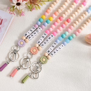 Pode incluir: Cordões de contas coloridas com os nomes "Mandy", "Norma" e "Sally" escritos em contas de letras brancas. Cada cordão apresenta um pingente de flor, uma borla e um porta-chaves. As contas são em cores pastel.