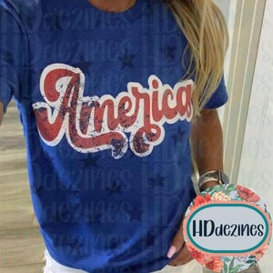 May include: A blue t-shirt with the word "America" in red and white distressed lettering. The shirt has a crew neck and short sleeves. Small blue stars are scattered across the shirt.