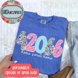 May include: A blue long-sleeve shirt with the numbers "42066" appliqued on the front. The numbers are in floral, pink gingham, and blue striped patterns. The words "home sweet home" are below the numbers. The shirt is on a white surface.