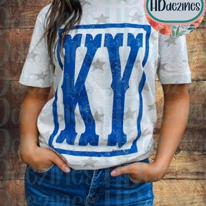 May include: A light-colored t-shirt with a blue "KY" graphic. The letters are outlined in a distressed style, with a star pattern on the shirt. The shirt has short sleeves and a crew neck.