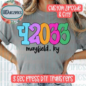 May include: A grey t-shirt featuring a large, colorful graphic with the number 42066 in blue, purple, orange, pink, and green. Below the number, the text reads mayfield, ky in black. The design is printed on a short-sleeve, crew-neck garment.