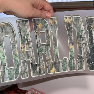 Op de afbeelding: Een transparante folie met het woord "DARLIN'" uitgesneden in een camouflagepatroon. De letters zijn wit omlijnd. Het camouflageontwerp heeft tinten groen, bruin en beige, die lijken op een boslandschap.