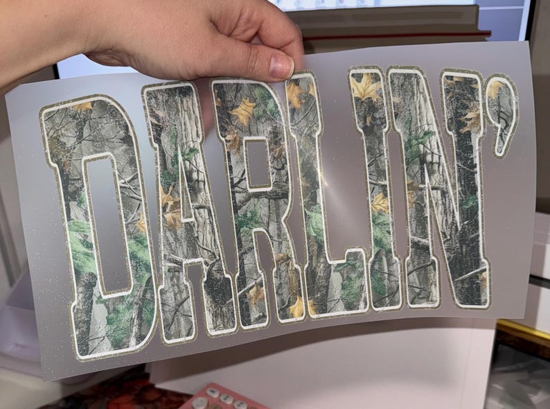 Op de afbeelding: Een transparante folie met het woord "DARLIN'" uitgesneden in een camouflagepatroon. De letters zijn wit omlijnd. Het camouflageontwerp heeft tinten groen, bruin en beige, die lijken op een boslandschap.